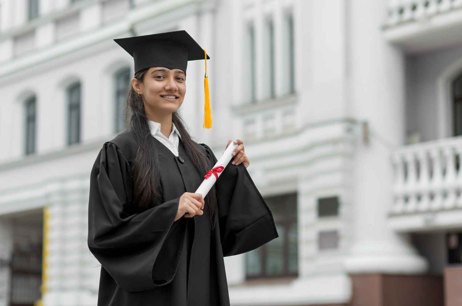 A smiling female graduate in a cap and gown holding a postgraduate degree, representing success through distance education courses, standing in front of a university building.