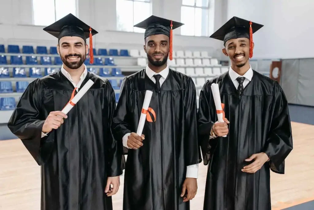 Three male graduates in black gowns and caps holding degrees, celebrating their success through distance education courses inside an auditorium.